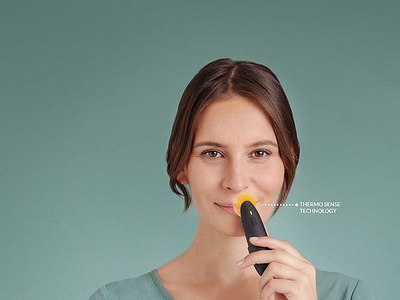 Eine Frau hält ein kleines schwarz-gelbes Gerät an ihre Oberlippe. Auf dem Gerät zeigt der Text „THERMO SENSE TECHNOLOGY“ vor einem einfarbig blaugrünen Hintergrund.