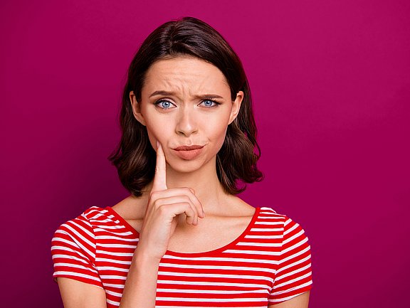 Eine Frau in einem rot-weiß gestreiften Hemd, die ihr Gesicht berührt und vor einem magentafarbenen Hintergrund verwirrt dreinschaut.