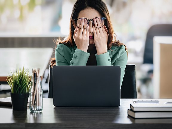Eine Frau sitzt an einem Schreibtisch mit einem Laptop, verdeckt ihre Augen mit den Händen und wirkt gestresst oder müde.