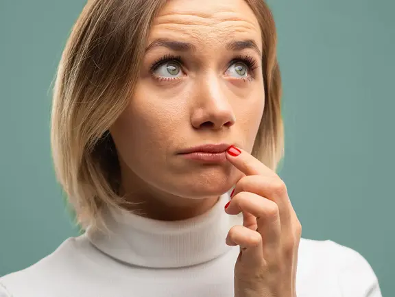 Eine Frau mit kurzen blonden Haaren und einem weißen Rollkragenpullover blickt nachdenklich auf und berührt mit dem Finger ihre Lippen.